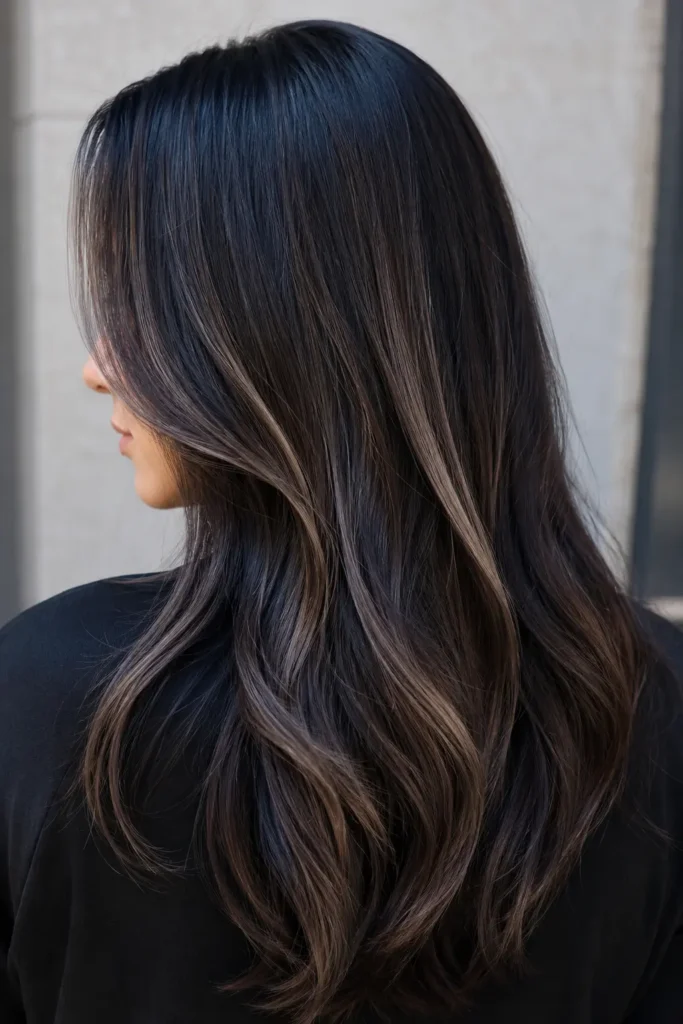 woman shown mostly from the back with a slight side profile, featuring sleek black hair enhanced with elegant ash brown highlights. Her silky, glossy, healthy hair is illuminated by bright natural daylight against a soft gray-beige background, with the hairstyle filling most of the DSLR-quality image.