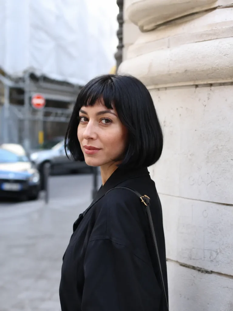 woman with a sleek jet-black bob haircut and short baby bangs, standing outdoors in an urban setting. She wears a trendy dark outfit and looks over her shoulder toward the camera.