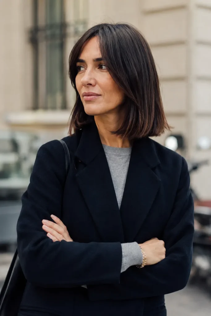 woman aged 30–50 with a sleek lob haircut and soft curtain bangs, featuring glossy dark brunette hair. She is wearing an elegant business-casual outfit and standing in an outdoor urban setting