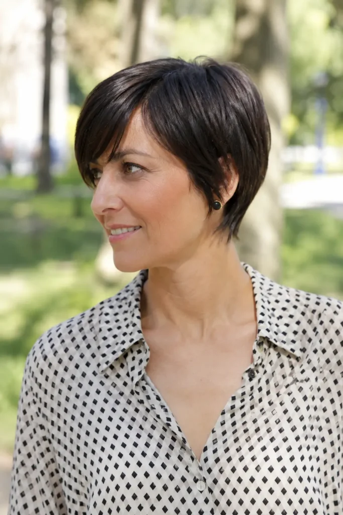 woman aged 30–50 with a sleek pixie bob haircut and side fringe, featuring glossy dark brunette hair. She wears an elegant patterned blouse and is photographed outdoors in a city park with a softly blurred green background