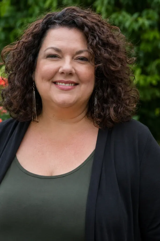 Plus-size woman aged 30–55 with soft layered curly rich brown hair standing in a lush outdoor garden. She wears a stylish casual outfit and smiles warmly at the camera. Natural skin texture and realistic facial features are visible in the soft daylight,