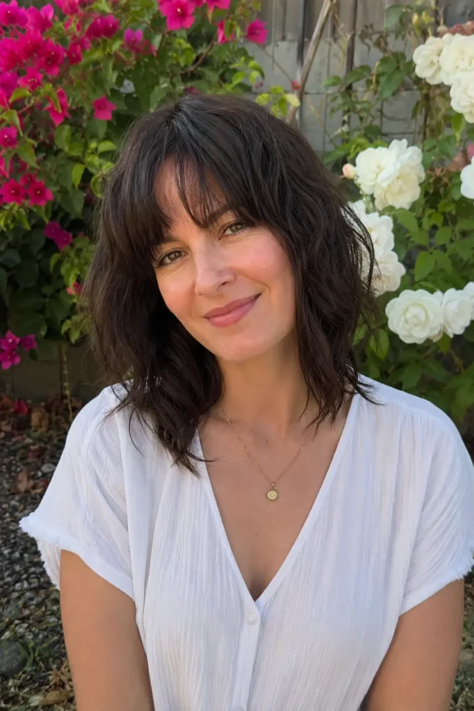 woman with a soft shag haircut and curtain bangs, textured brunette hair, and natural skin texture, wearing a white casual blouse in an outdoor floral garden. The upper-body portrait highlights the layered hairstyle