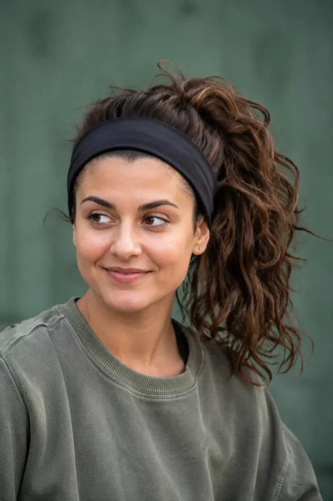 25-year-old woman wearing a black sporty headband with a voluminous high ponytail hairstyle. Her dark brown hair has natural texture, movement, and fullness, making the hairstyle the focal point of the portrait.