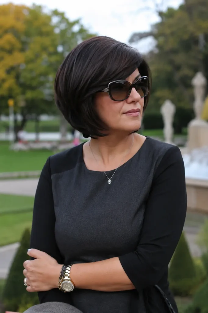 woman aged 30–50 with a stacked bob haircut and side-swept bangs, smooth dark brunette hair, and realistic skin texture, wearing an elegant dark outfit in an outdoor park setting