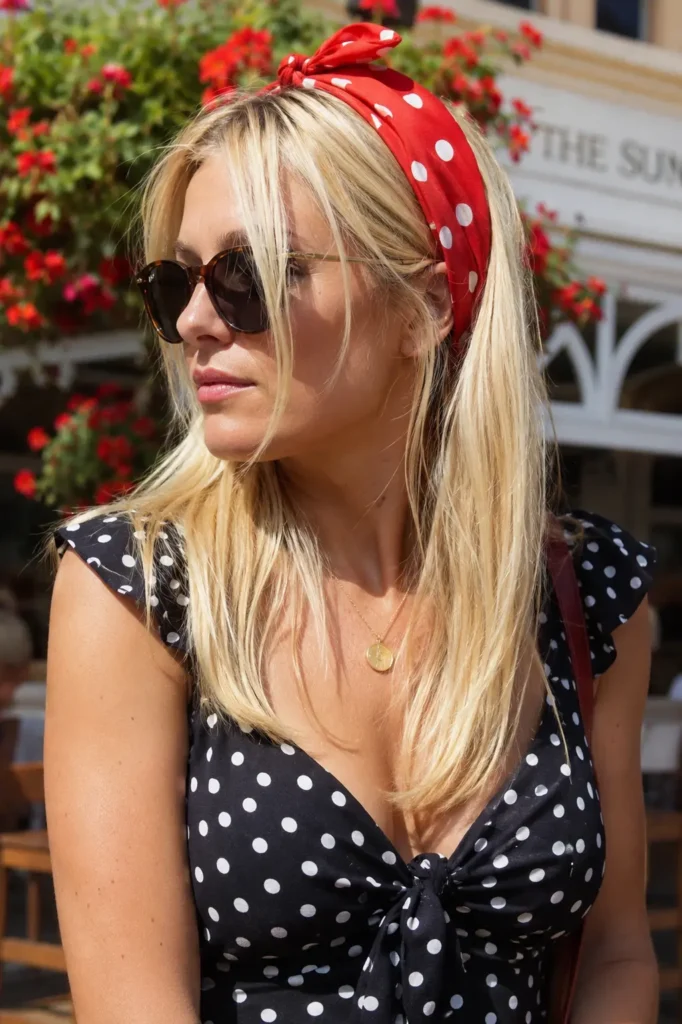 English woman with sleek straight blonde hair wearing a red polka-dot bandana headband, photographed outdoors in natural sunlight.
