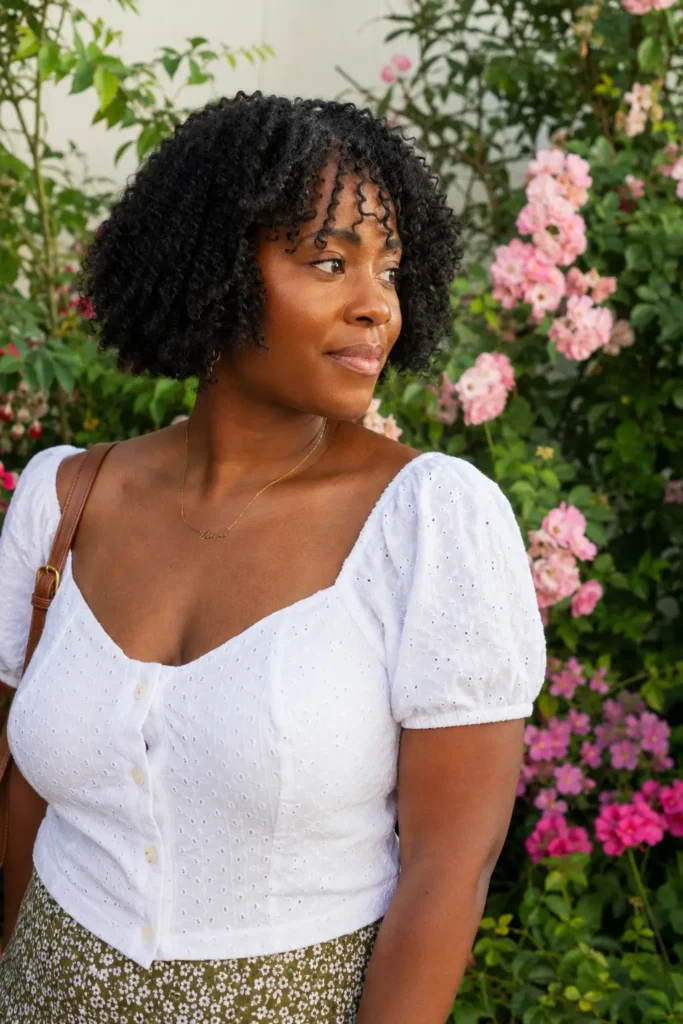 real Black woman with a textured natural bob haircut and curly bangs, featuring defined natural coils and healthy volume. She wears a casual white blouse and stands in a blooming garden filled with pink flowers