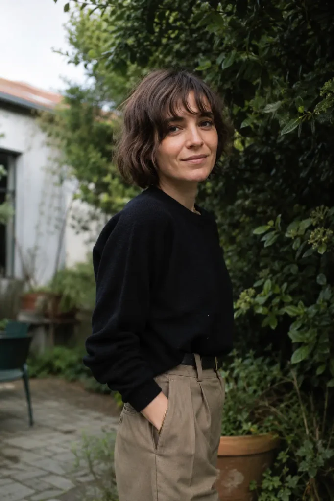 woman with a tousled bob haircut and piecey bangs, featuring medium brown hair and natural texture, posing outdoors in a lush garden setting. She wears a casual chic outfit and has realistic skin texture and authentic facial features