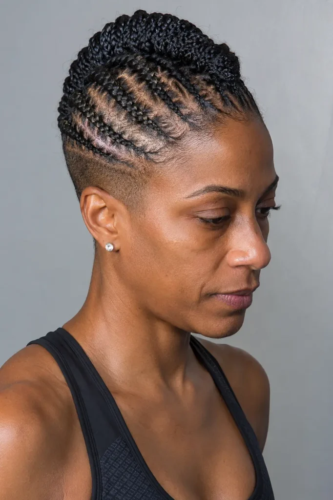 woman with a mini braid hawk hairstyle featuring multiple small, neatly braided cornrows leading into a raised braided center section. The sides are closely cropped, highlighting the detailed braid pattern and scalp design. She wears a black sleeveless athletic top