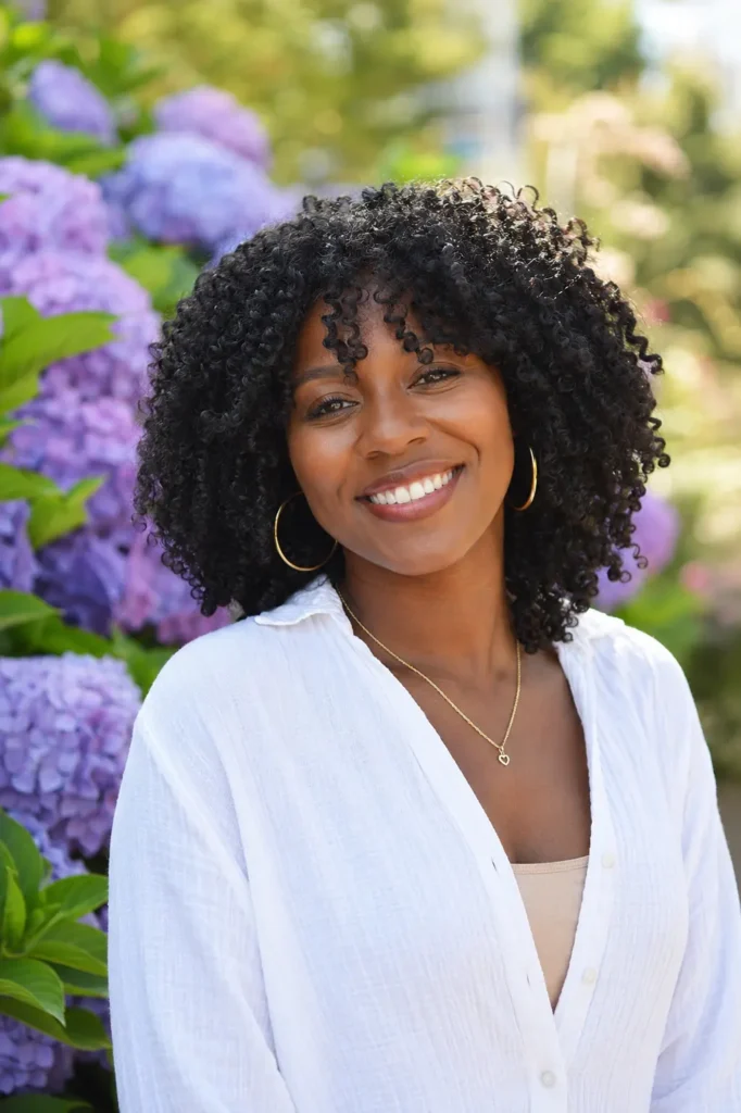 Black woman with a wash-and-go hairstyle featuring defined natural curls, smiling in an outdoor floral garden. Her healthy textured hair is prominently displayed, with realistic skin texture and a casual chic outfit