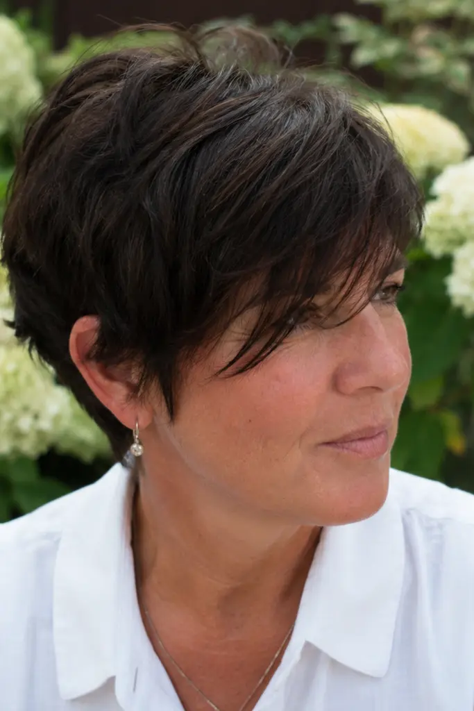 Woman over 40 with a wispy brunette pixie haircut and long side-swept bangs, photographed outdoors in natural summer daylight