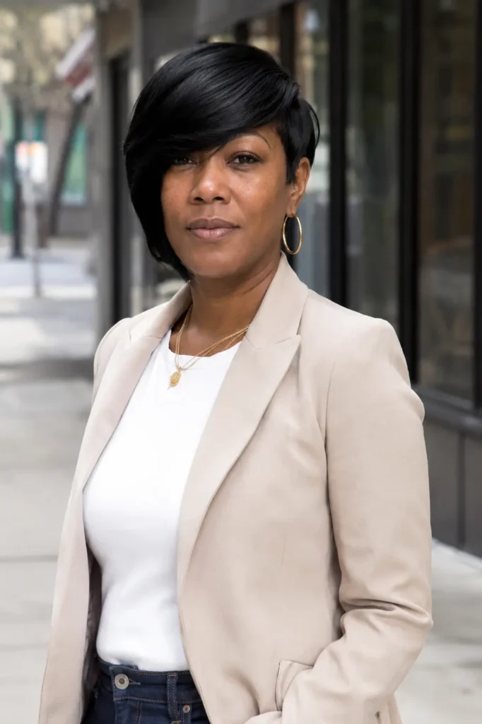 Black woman with a sleek asymmetrical bob haircut and side-swept bangs, standing outdoors in an urban setting. She wears a stylish beige business-casual blazer over a white top, with glossy black hair framing her face