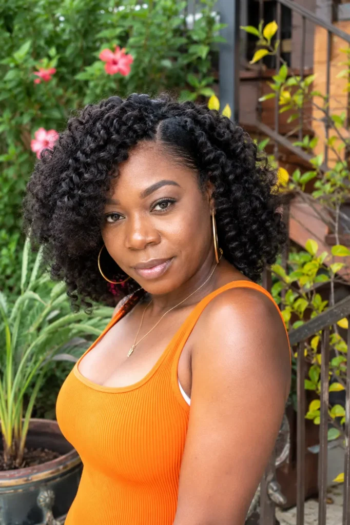 Real Black woman with a curly Bantu knot-out hairstyle featuring voluminous, defined curls standing in an outdoor garden setting. She wears a stylish orange sleeveless outfit and gold hoop earrings