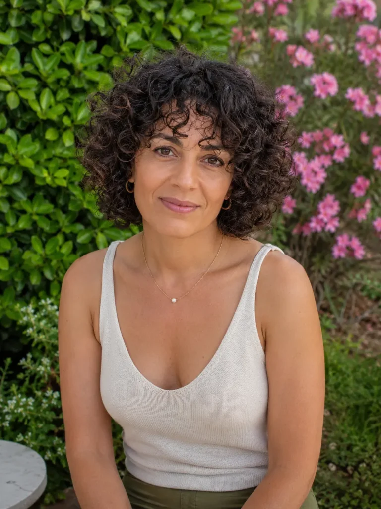 woman with a curly bob hairstyle and curly fringe, featuring defined natural dark curls and realistic skin texture, wearing a casual elegant sleeveless top while sitting in an outdoor garden with blooming pink flowers and lush greenery