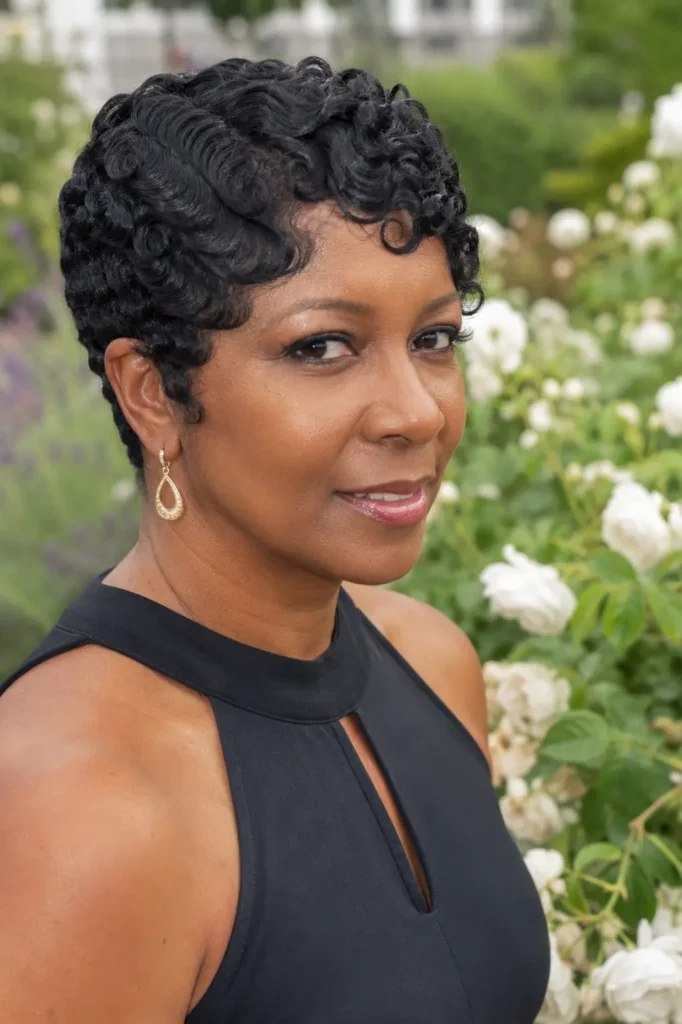 Black woman aged 30–55 wearing an elegant sleeveless black outfit, standing in a blooming outdoor garden. She has a curly finger wave pixie hairstyle with sculpted waves and defined curls that frame her face beautifully
