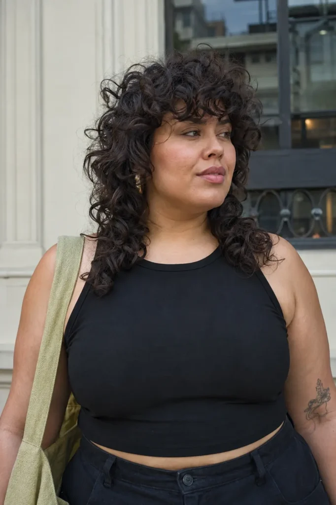 Close-up portrait of a real plus-size woman aged 25–45 with a curly wolf cut hairstyle featuring layered, textured curls and natural volume. The defined curls frame her face and showcase the haircut's shaggy shape and movement.