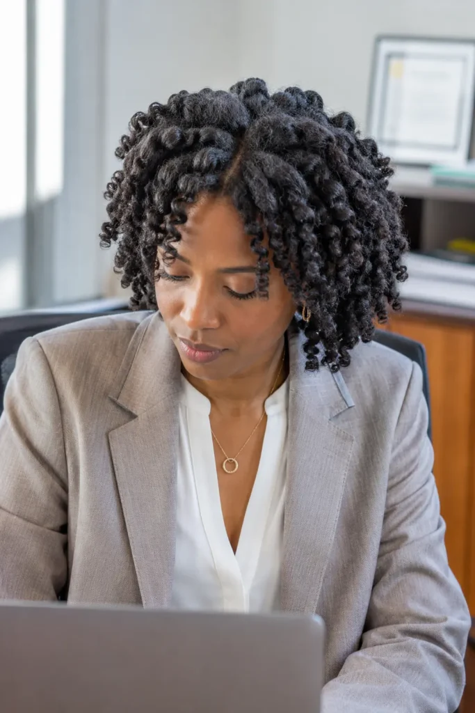 woman with a defined twist-out hairstyle on natural Type 4 hair, showcasing detailed curl definition and natural volume. She is seated in a professional office setting, wearing business-casual attire and working at a laptop