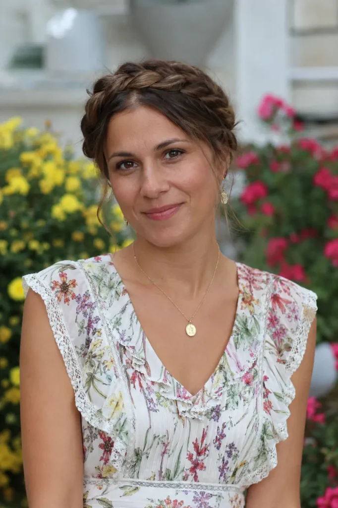 woman aged 25–40 with a halo braid encircling her head, warm brunette hair, and natural beauty, wearing an elegant floral dress in a colorful outdoor garden.