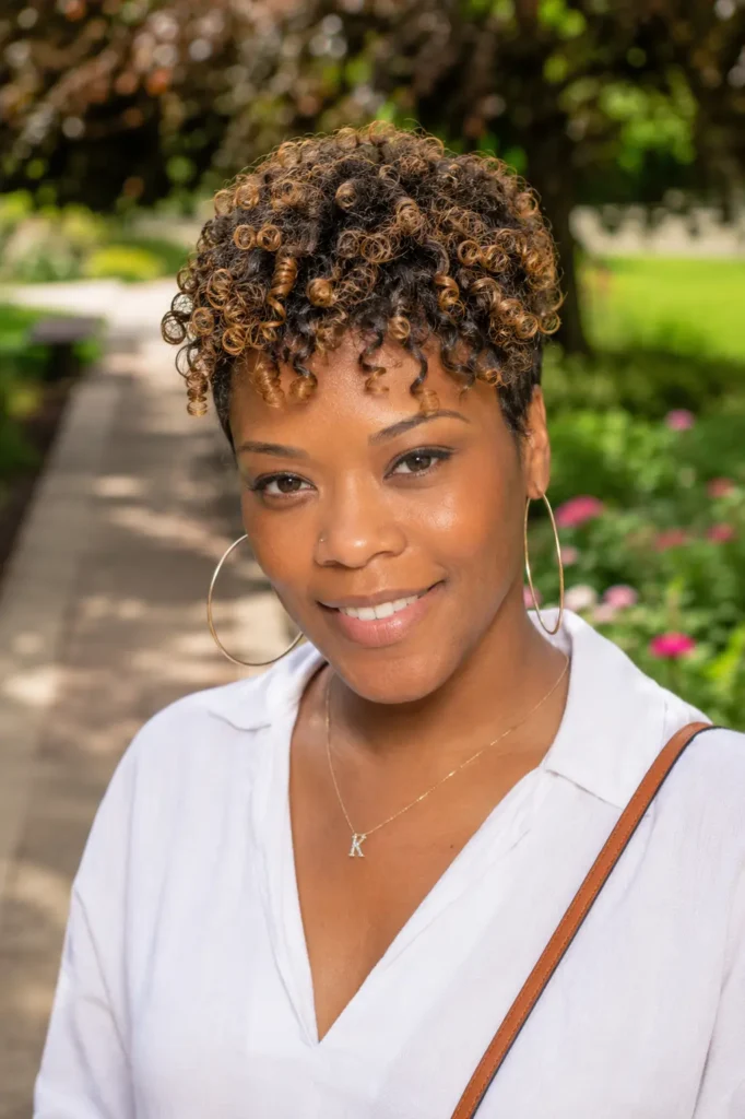 Black woman aged 25–45 with a highlighted curly crop hairstyle featuring honey-blonde accents. Her defined natural curls are styled short and voluminous on top, showcasing texture and dimension. She wears a chic casual outfit and smiles warmly while standing in a lush outdoor garden