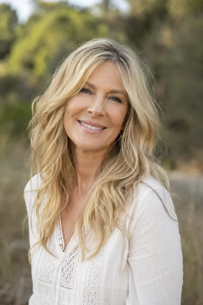 Mature woman aged 55–65 with long champagne blonde wavy hair, smiling outdoors in soft daylight. She wears an elegant casual white blouse