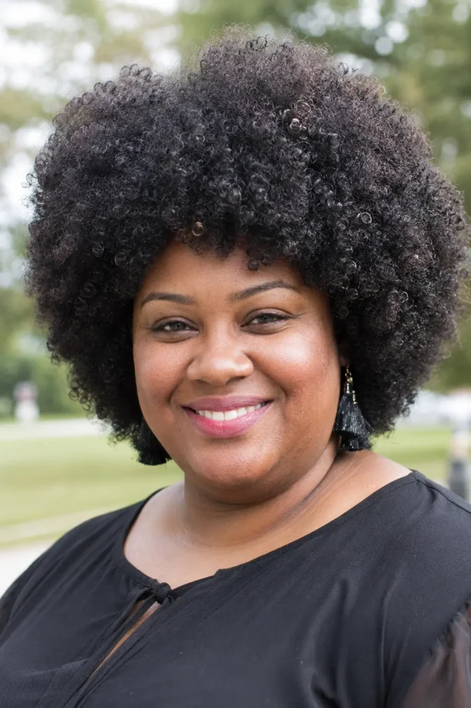 Close-up portrait of a plus-size woman with a perfectly rounded curly afro hairstyle, featuring dense natural curls and a full, balanced shape. The hairstyle fills most of the frame, with detailed curl texture in sharp focus