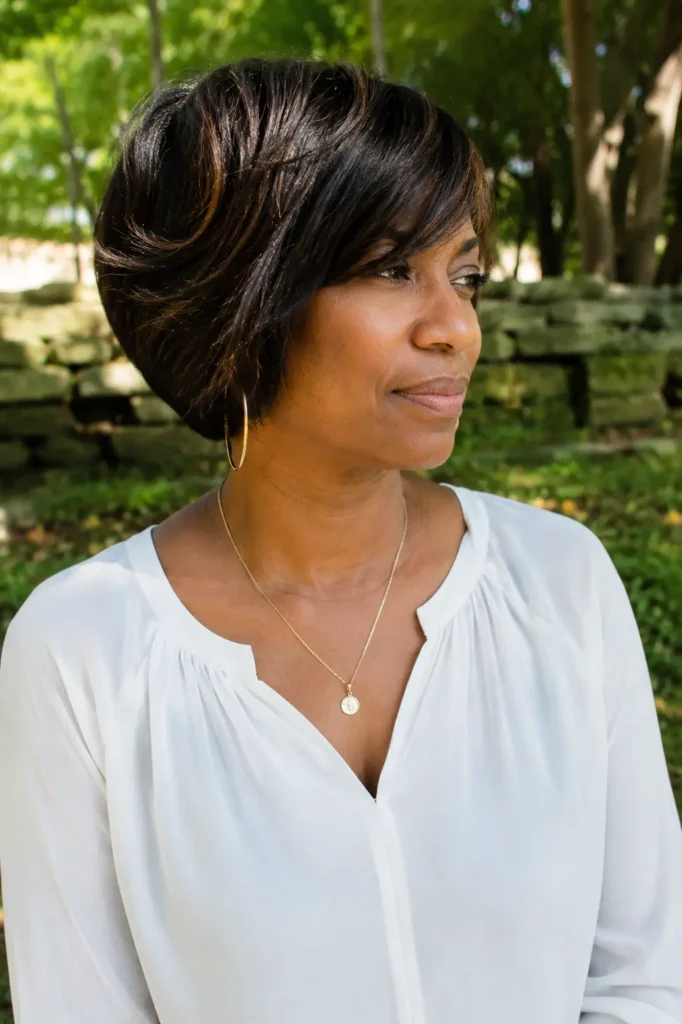 Black woman aged 35–55 with a stacked bob haircut and side-swept bangs, smooth dark brunette hair with subtle shine, standing in a lush outdoor park. She wears an elegant white blouse and delicate jewelry