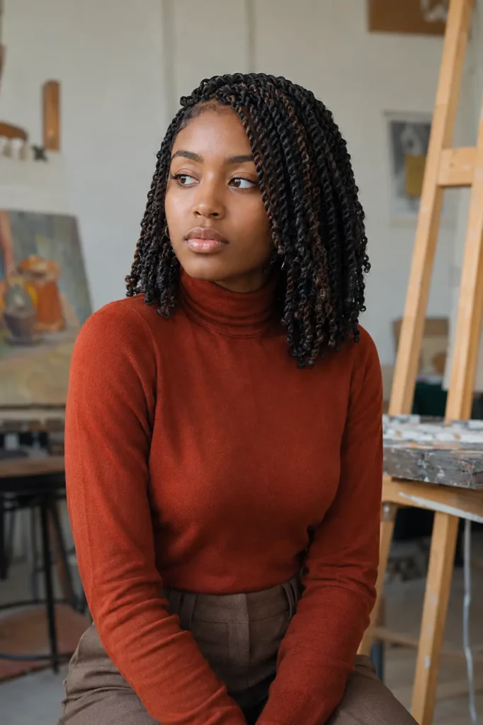 a 20-year-old Black woman with shoulder-length Senegalese twists featuring subtle dark-brown highlights. She wears a rust-orange turtleneck and tailored brown trousers in a minimalist art classroom with softly blurred easels and paintings