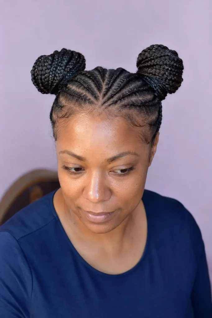 Black English woman wearing symmetrical cornrow braids styled into two neat space buns, photographed from a slightly elevated front angle with a pale lavender background and natural daylight.