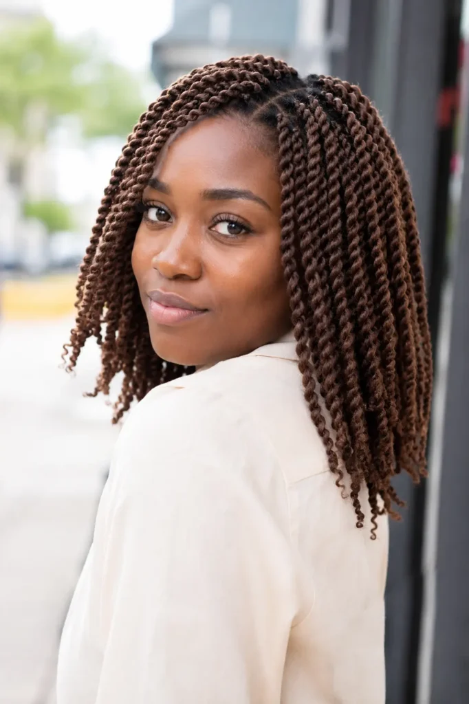 a Black woman with shoulder-length chestnut brown twist braids, wearing a cream blouse in modern English fashion, captured in bright natural daylight with realistic skin texture and the hairstyle as the main focus.