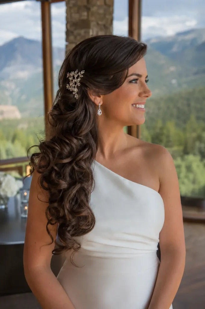 white bride with long rich brunette side-swept bridal curls, adorned with a crystal hair comb, wearing a one-shoulder satin wedding gown at a bright mountain lodge wedding venue.