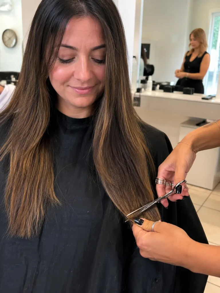 a woman with long healthy hair sitting in a modern salon while a hairstylist trims the split ends with professional scissors