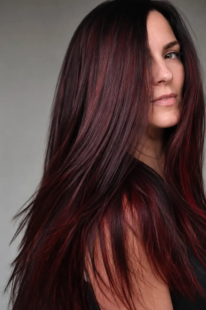 white woman with very long sleek black cherry hair featuring deep burgundy, ruby, and cherry-red reflections, styled straight with a glossy finish against a neutral minimalist background.