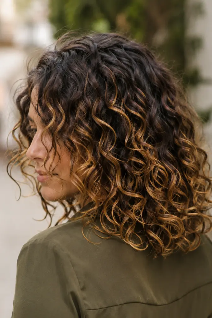 a woman with shoulder-length defined curls featuring a seamless dark-to-light ombré from natural roots to warm caramel ends, wearing a deep olive-green top outdoors in soft afternoon daylight