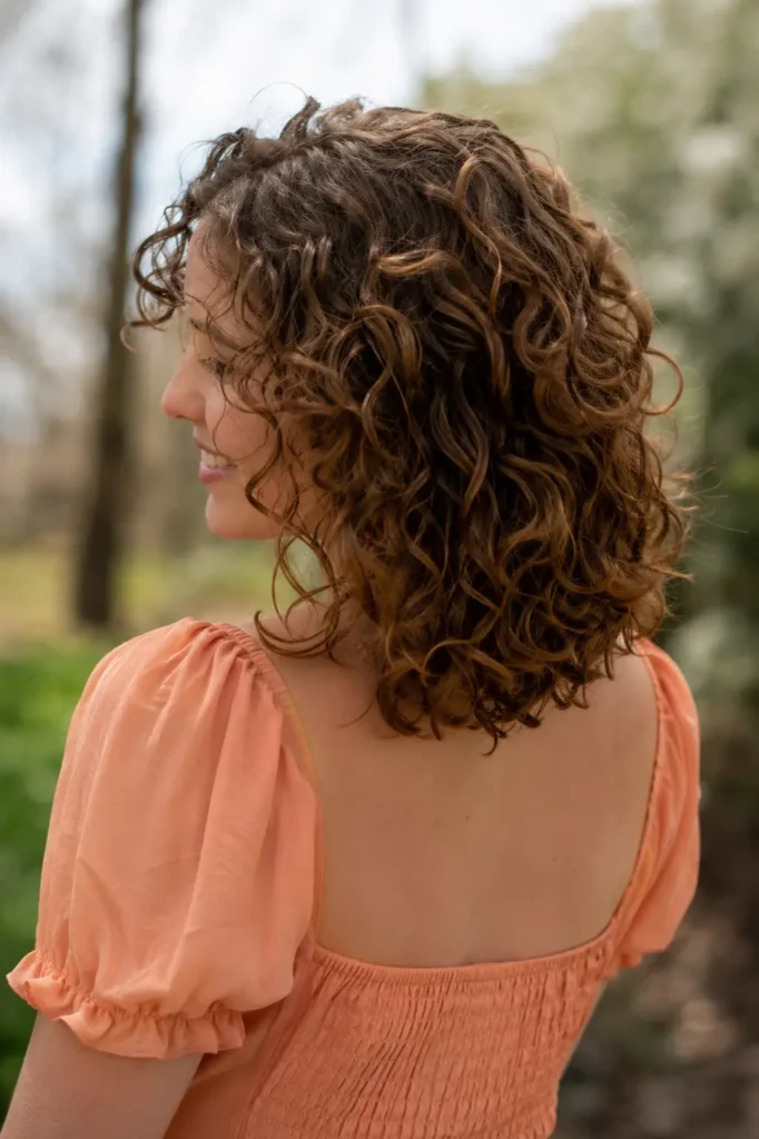 white woman in her late 20s with shoulder-length natural brown curls, shown from a three-quarter rear view, wearing a soft peach dress outdoors in gentle natural daylight, with the curls as the main focus.