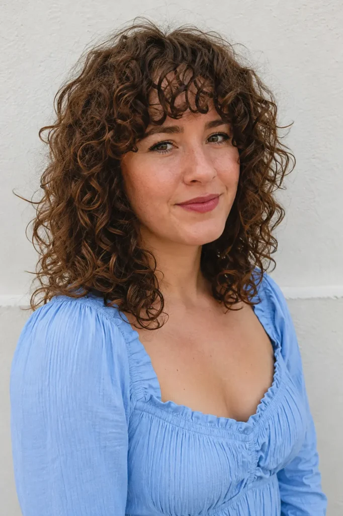 white woman with shoulder-length curly hair and short curly fringe, wearing a sky-blue dress in natural light.