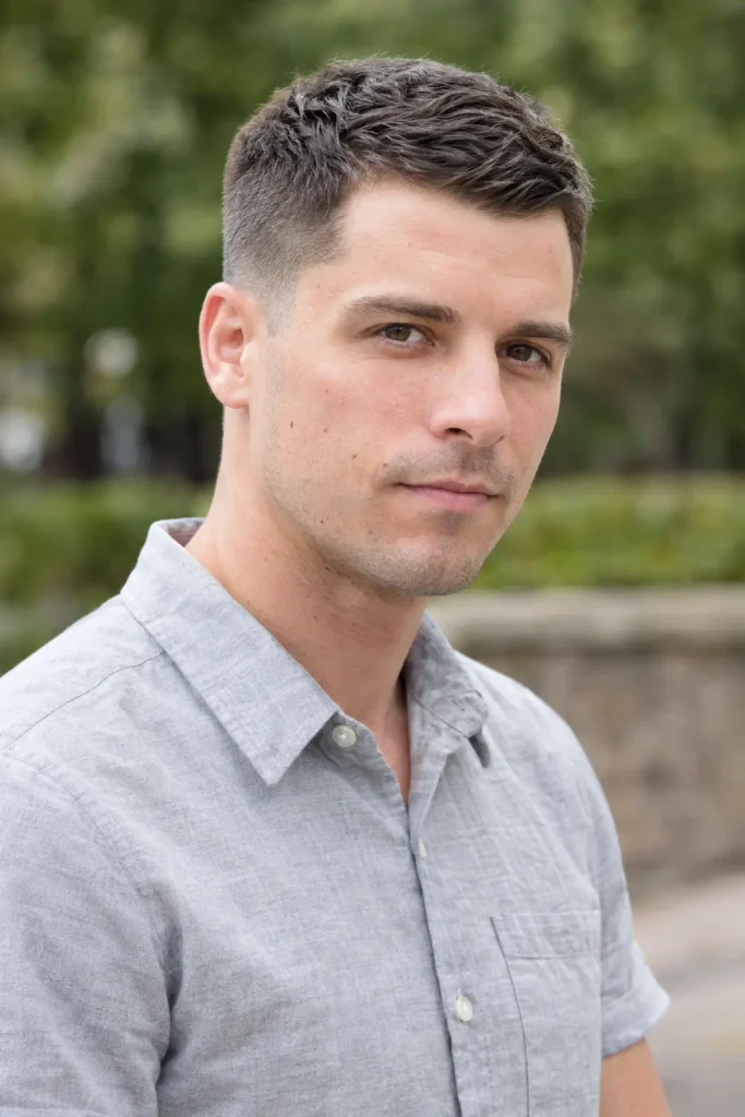 Real young white man with a classic tapered crew cut and short textured top, wearing a light-gray button-up shirt outdoors.