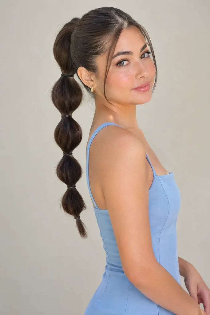 young woman with a sleek bubble ponytail featuring multiple rounded sections, wearing a light blue sleeveless mini dress in a neutral setting.