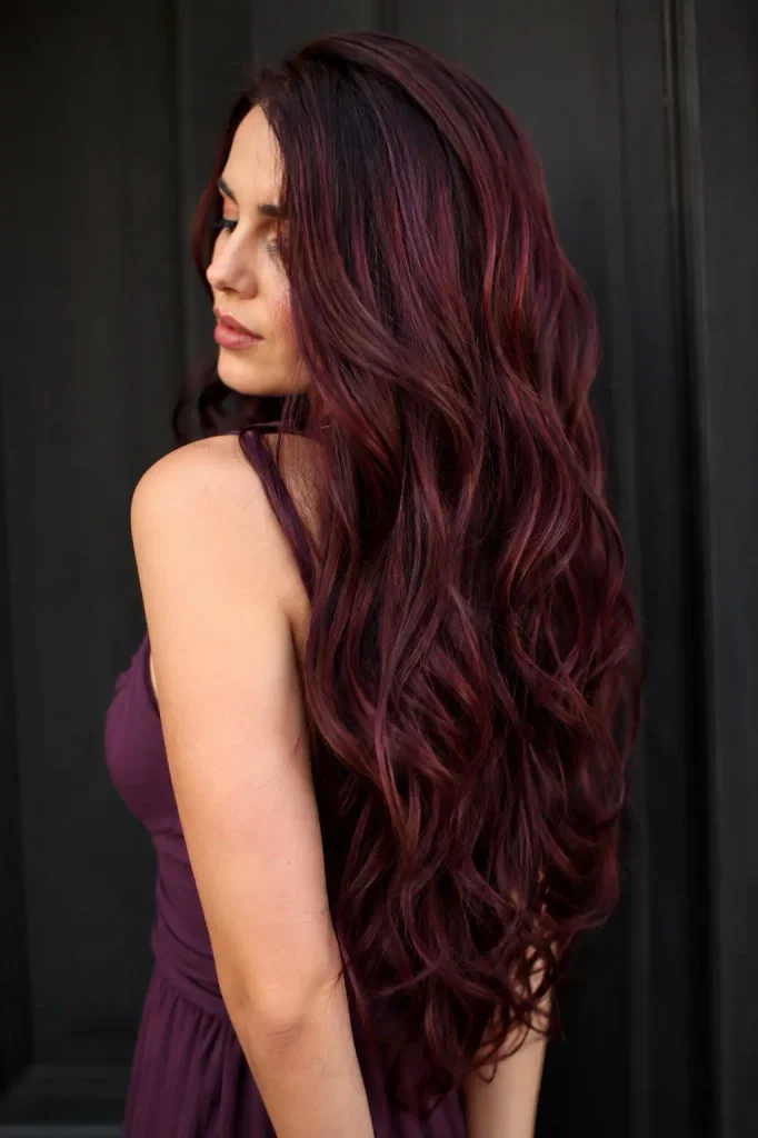 a real adult woman with extremely long, thick burgundy wine-colored wavy hair, showing rich plum and violet undertones, glossy dimensional color, and natural hair texture. She wears a fitted purple dress against a dark neutral background.