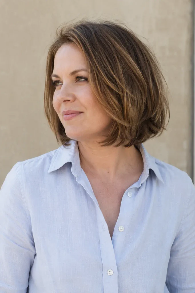 Chin-length layered bob haircut on a woman with a round face, featuring soft layers below the cheekbones, a slightly off-center part, and natural movement through the ends, paired with a pale blue linen shirt.
