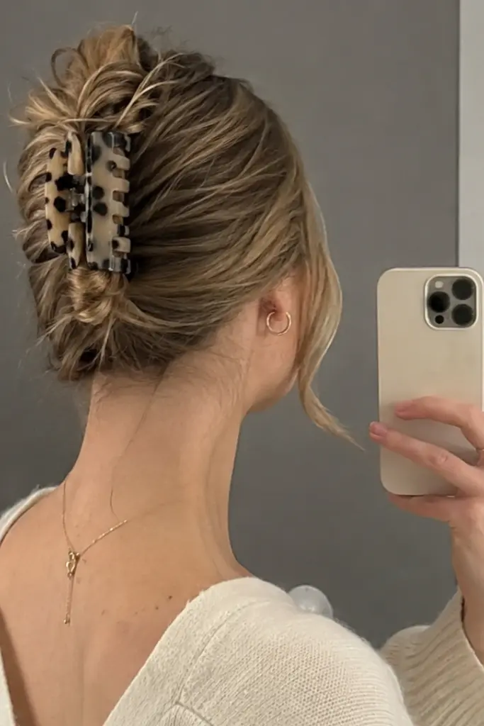 Photorealistic mirror selfie of a 23-year-old white woman wearing a claw clip French twist updo for medium-length hair, with soft layers, loose strands, and natural hair texture, dressed casually in a cream knit outfit.
