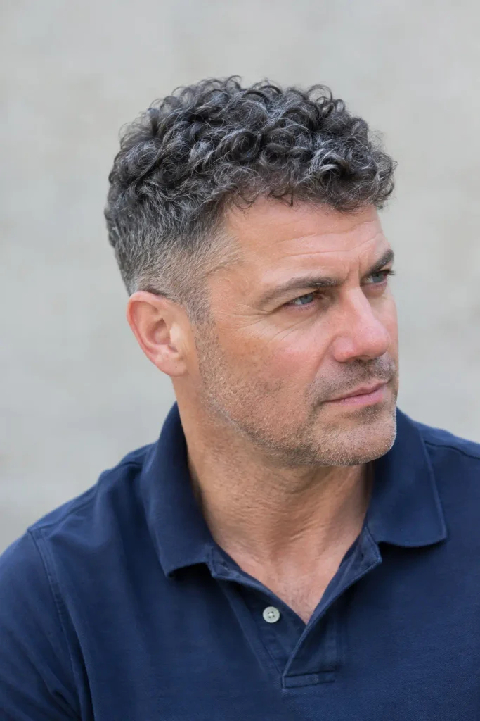 mature white man with a short textured salt-and-pepper curly crew cut, neatly tapered sides, wearing a dark blue polo shirt in natural daylight.