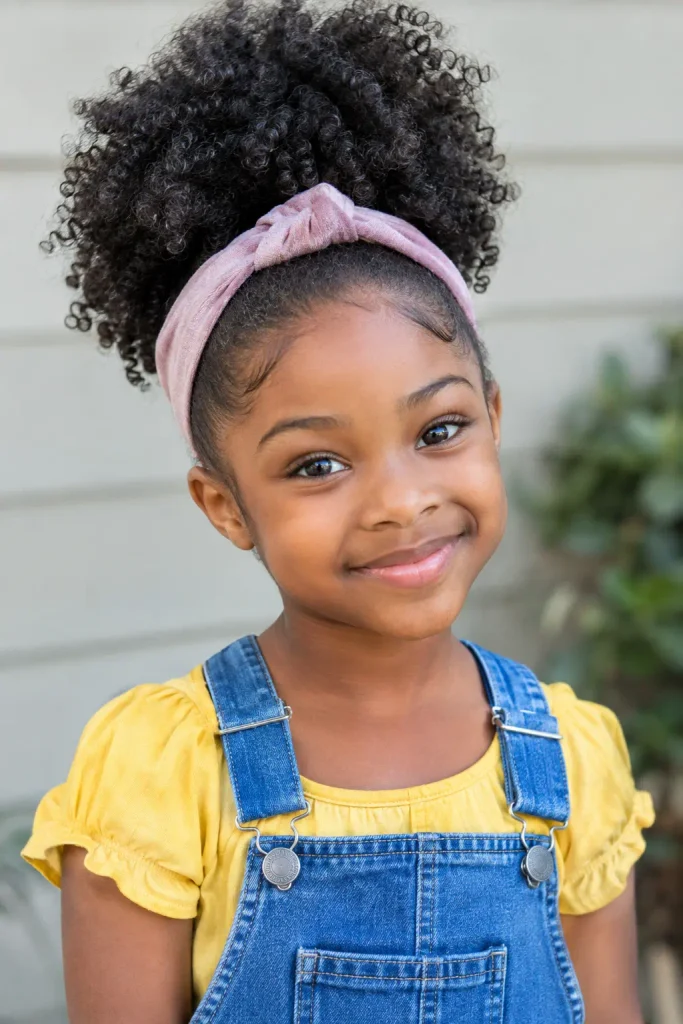 a Black young girl with naturally defined coily curls gathered into a full high puff, wearing a soft fabric headband, yellow cotton blouse, and denim overalls.