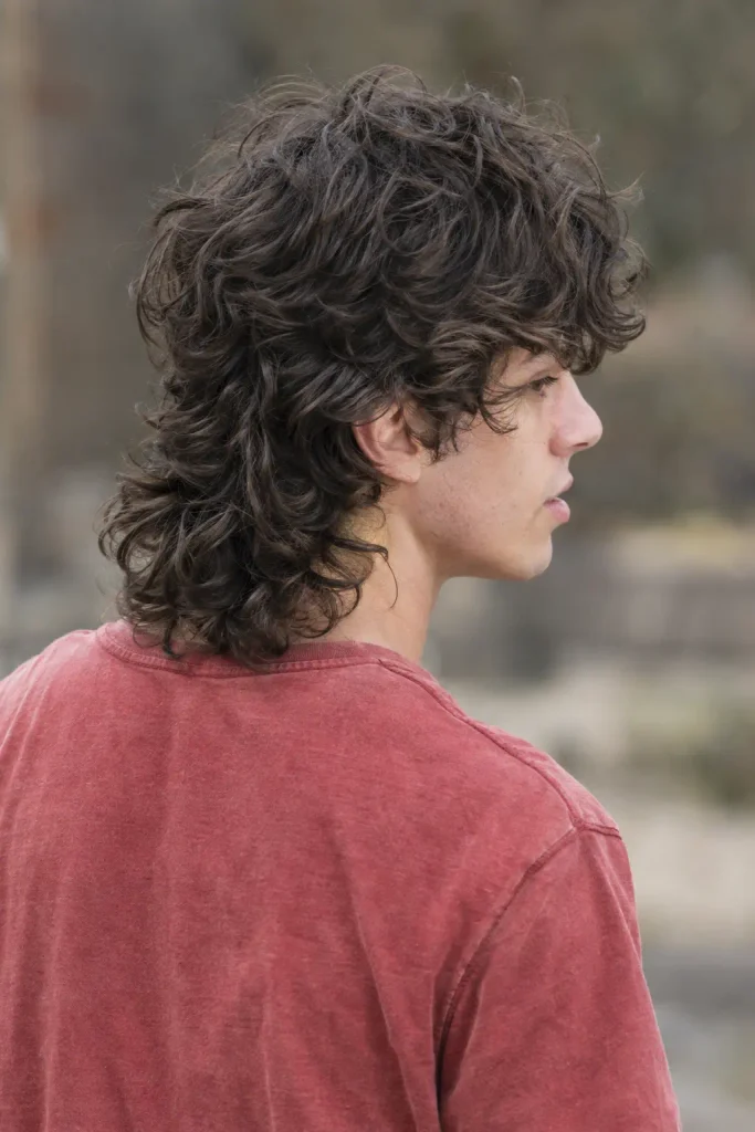 Young man with thick dark brown curly hair styled in a layered curly wolf cut with long textured curls around the neck, viewed from behind outdoors.
