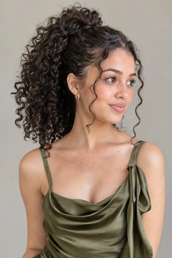 Young woman with a voluminous defined curly ponytail, natural separated curls, and soft tendrils, wearing a contemporary satin cocktail dress in a neutral setting.