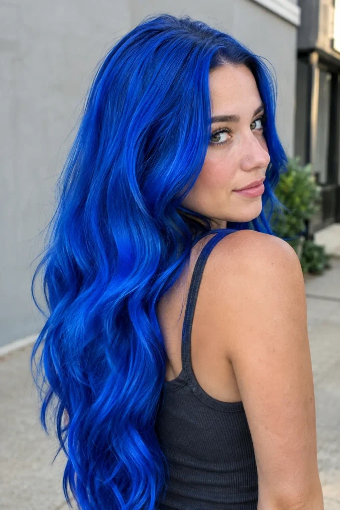 Young woman with long vivid cobalt-blue wavy hair photographed from a rear three-quarter view outdoors, wearing a charcoal sleeveless top.