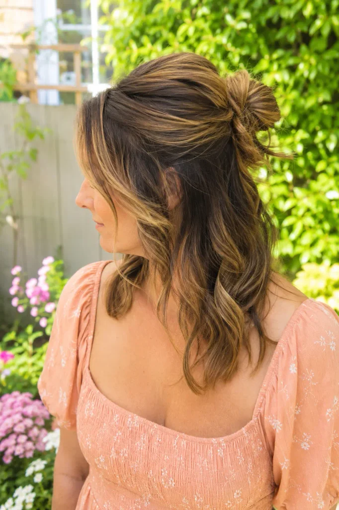 white woman with medium-length wavy hair styled in a textured half-up bun, wearing a peach Western-style dress in a bright English garden.