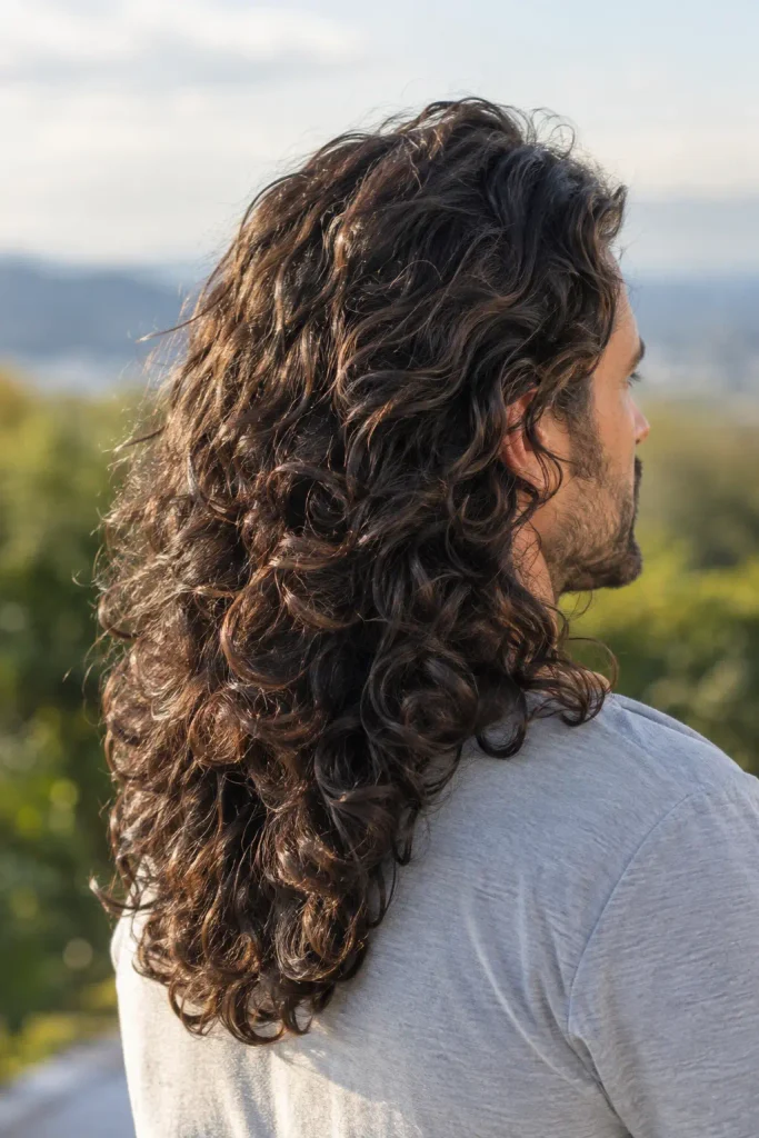 man with long, thick dark brown curly hair cascading past his shoulders, viewed from the back in warm outdoor daylight.