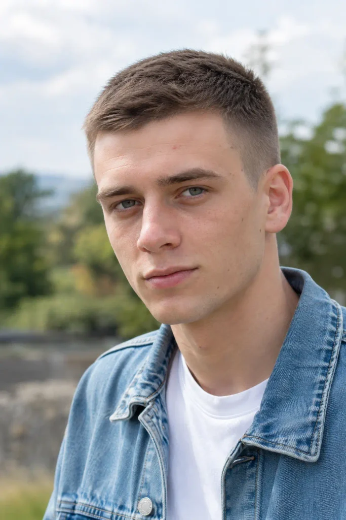 young white man wearing a denim jacket and white T-shirt, showing a very short crew cut with a compact textured top and clean tapered sides outdoors in natural daylight.