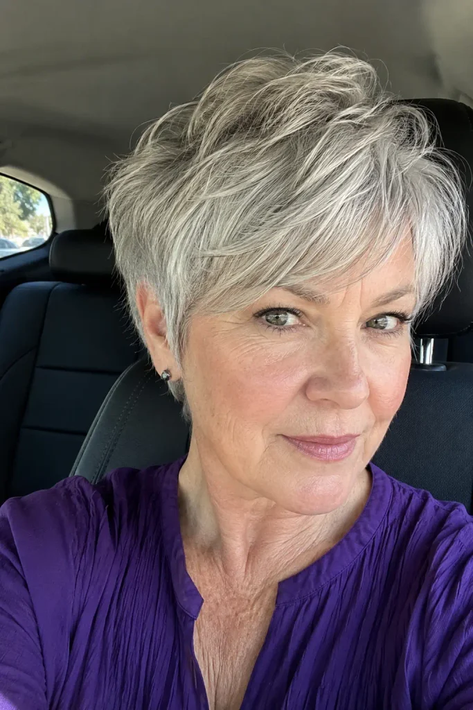62-year-old woman with fine silver-gray long pixie haircut, textured crown and soft sweeping fringe, wearing a purple dress inside a car in natural daylight.
