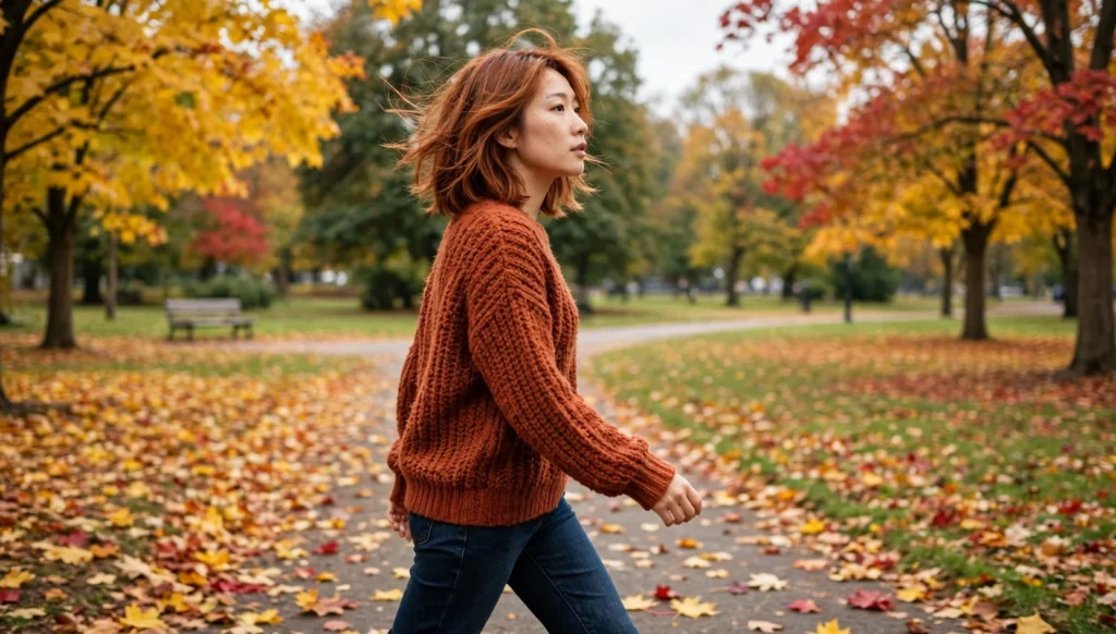 Young woman walking through an autumn park in a three-quarter view, wearing a rust sweater, with a copper layered fall haircut.