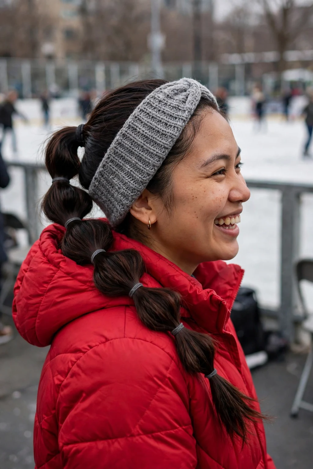 Young woman with a bubble braid ponytail and a knit headband, wearing a red coat at an ice rink.